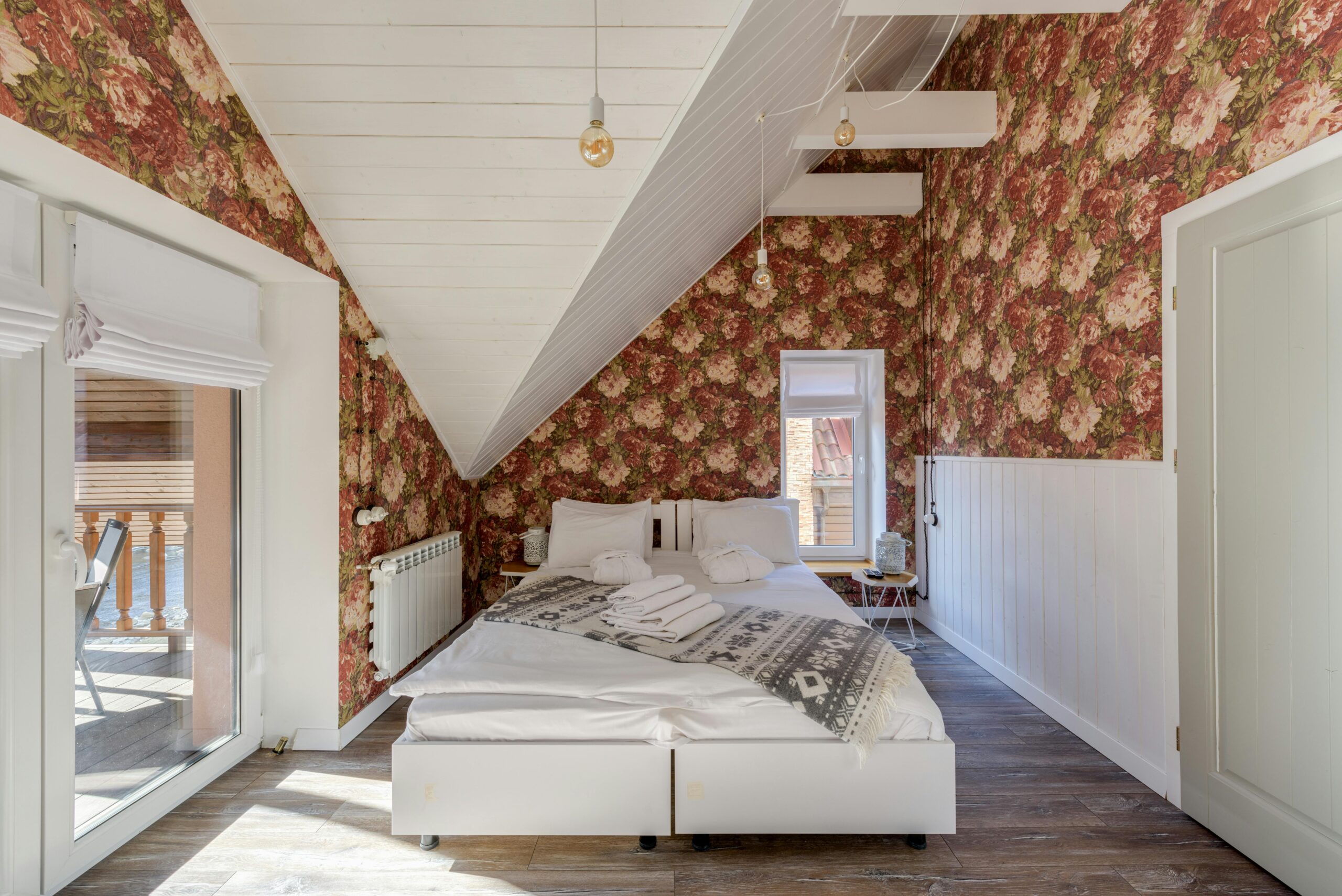 Bright attic bedroom with floral wallpaper, bed, and natural light from glass windows.